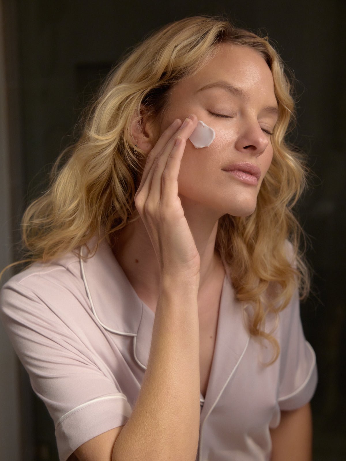 A person with long, wavy blonde hair applies a small amount of Cozy Earth's Night Cream to their cheek. Wearing a pale pink top with white piping, they have their eyes closed and appear relaxed against a softly blurred background.