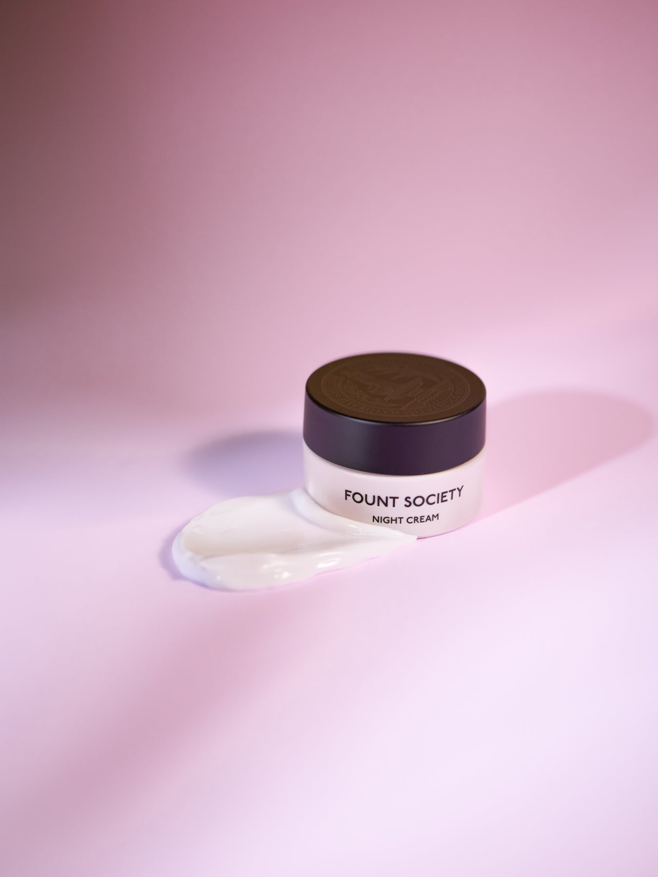 The Fount Society Night Cream mini container is surrounded by rich, luxurious cream. It appears luminous and glowing in warm purple tones. |Size:Mini