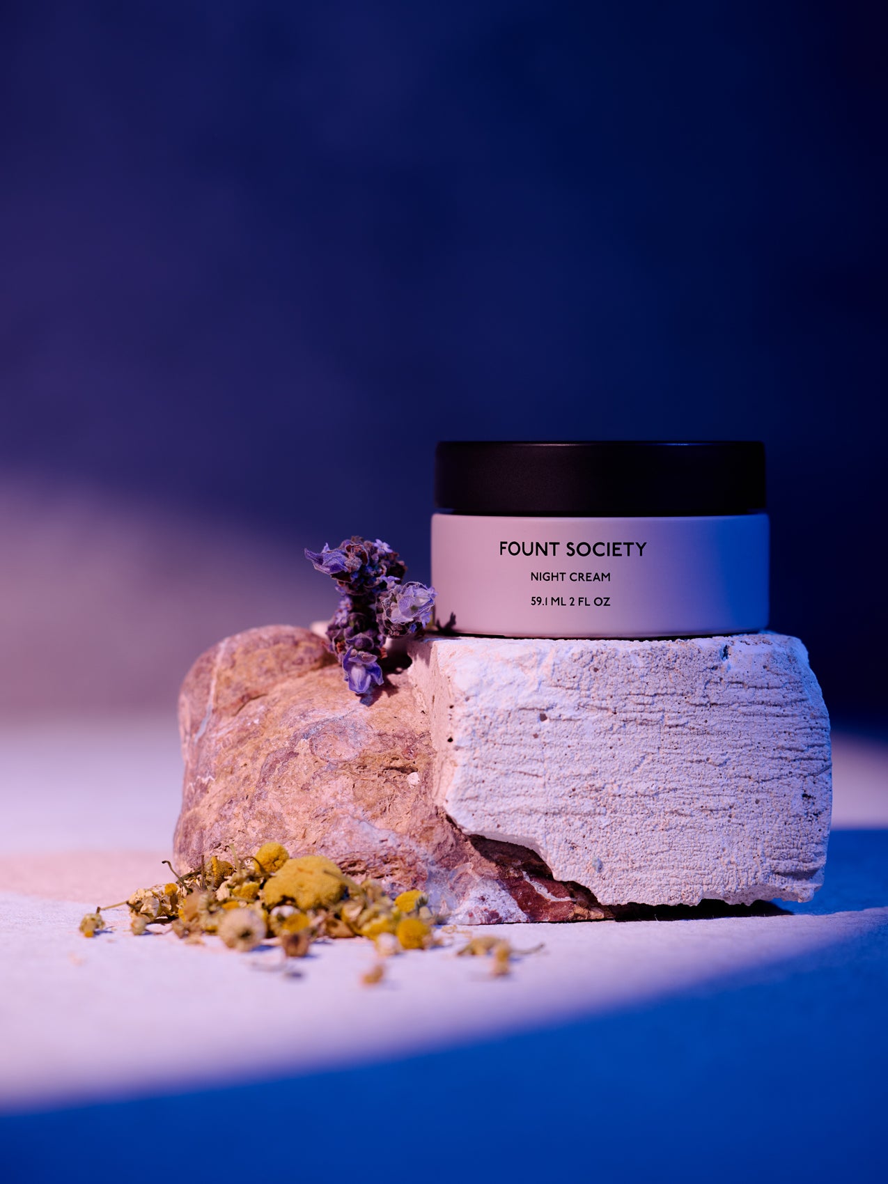 The Fount Society Night Cream container sits on a rough stone with dried flowers surrounding it. The lighting is moody in purple and deep blue tones. |Size:Full Size