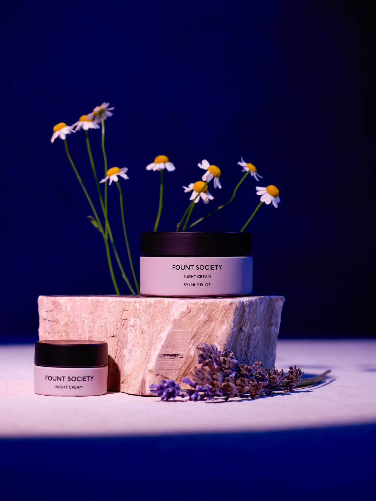 The Fount Society Night Cream in full size and mini size are pictured together on a stone with flowers surrounding them. |Size:Mini