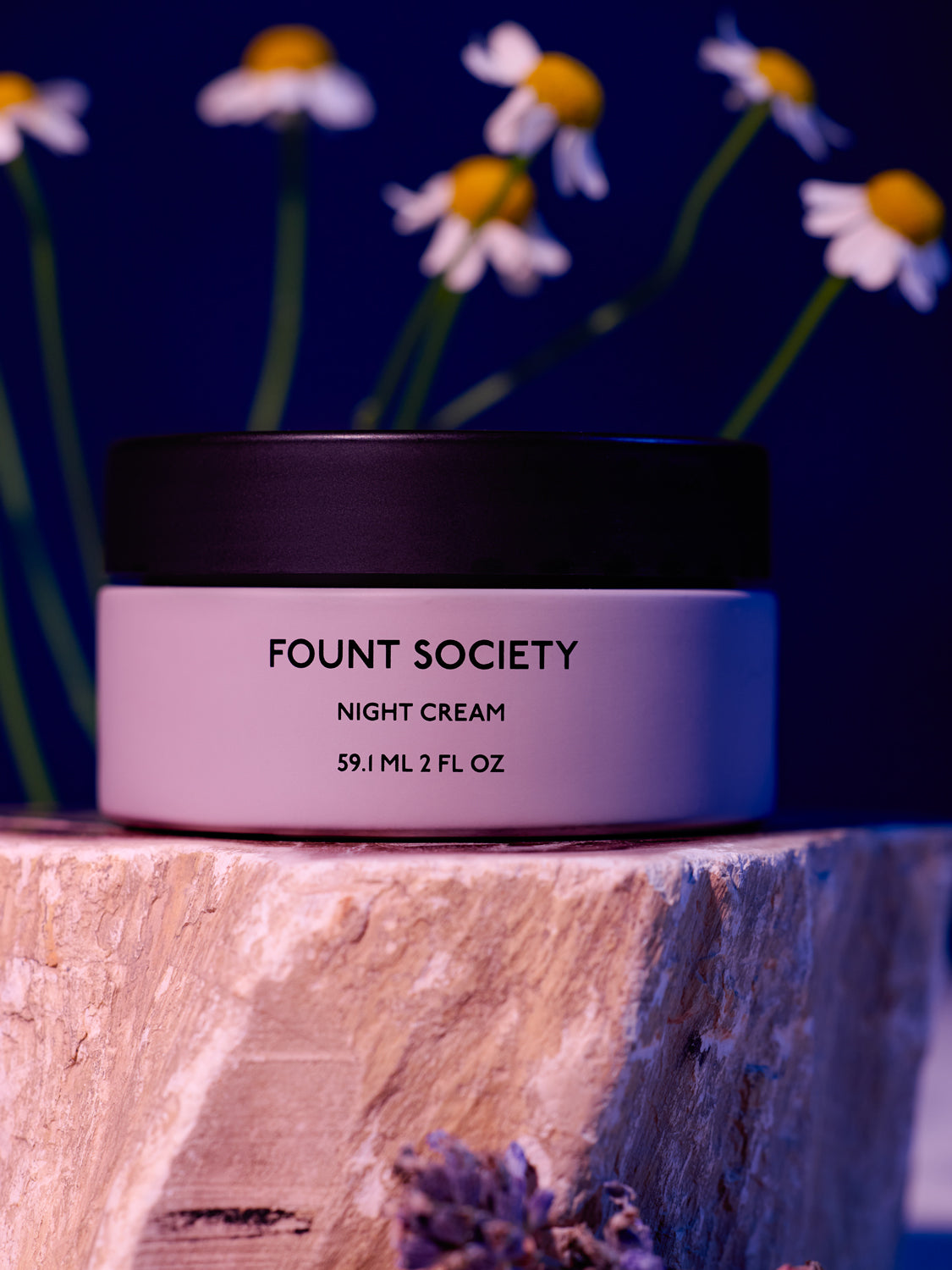 The Fount Society Night Cream container sits on a rough stone with flowers surrounding it. The lighting is moody in purple and deep blue tones. |Size:Full Size